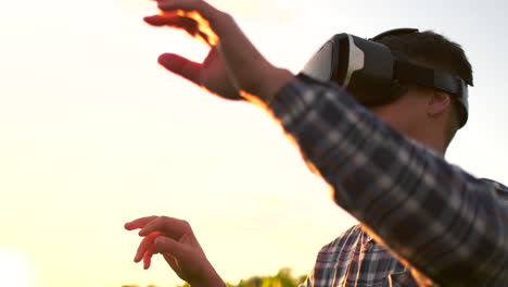 A-male-farmer-in-virtual-reality-glasses-wearing-a-plaid-shirt-and-jeans-in-a-field-at-sunset-controls-the-corn-irrigation-system.-Modern-farmer-new-technology-farmer-of-the-future